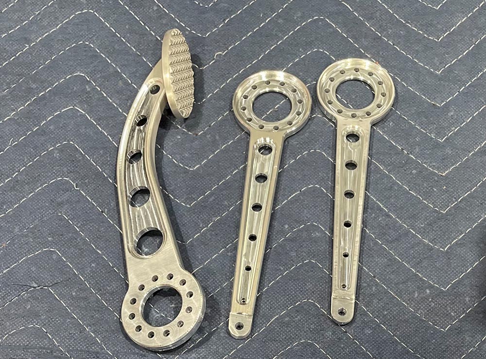 Three machined aluminum pedal parts on a gray quilted mat. One is a curved pedal with a textured oval foot pad; the others are straight linkage arms. All pieces feature circular lightening holes.