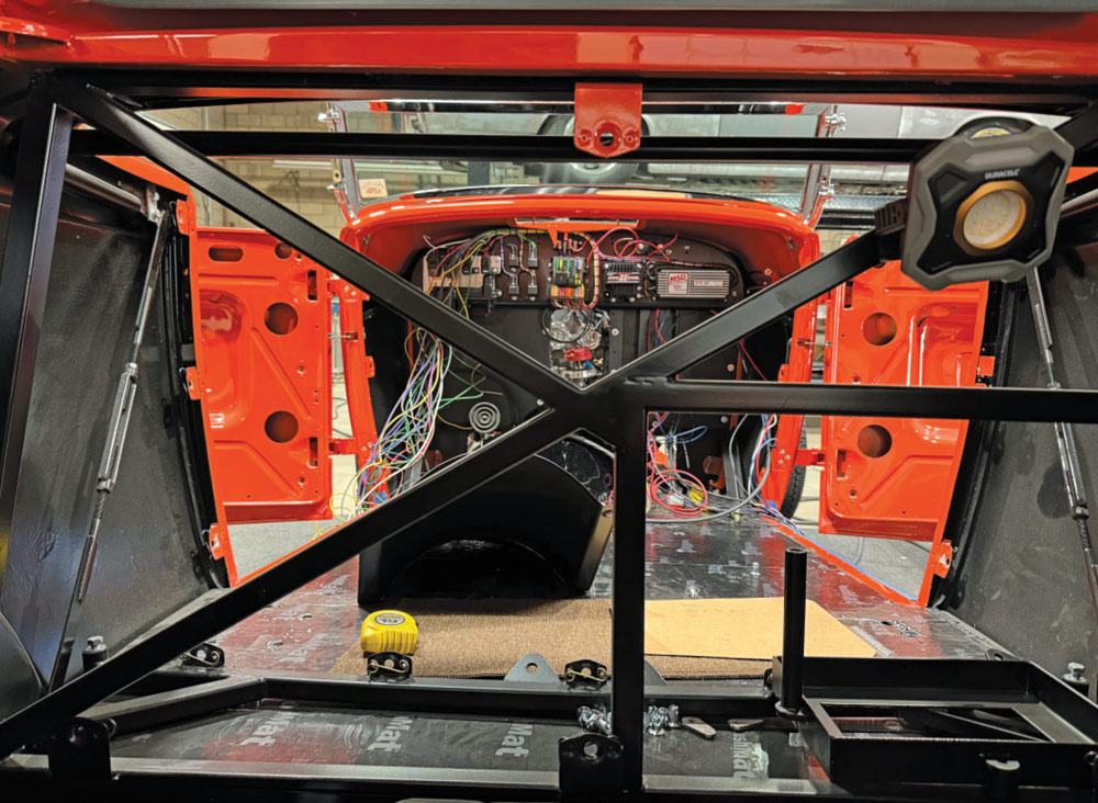 View from the trunk looking through a black metal X-brace into the red interior. Exposed wiring and electrical modules are visible on the firewall. The floor is covered in silver insulation with a yellow tape measure resting in the center.