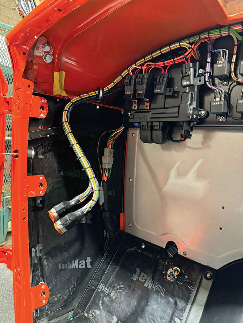 Close-up of the red roadster's interior footwell showing neatly routed wiring harnesses and connectors. Electronic modules are mounted to a silver panel, with black HushMat insulation lining the inner body panels.