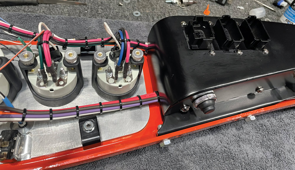 Rear view of the red dash panel showing intricate wiring from circular gauges connecting to a black fabricated cover. The cover features three large rectangular electrical ports to facilitate easy disconnection of the dash assembly.