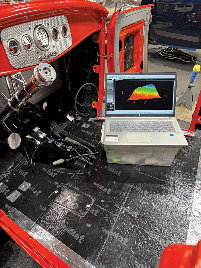 Interior view of the red roadster with a laptop displaying a colorful 3D tuning map in the driver’s footwell. The cabin features black HushMat flooring and a red dash with a polished, engine-turned gauge panel.