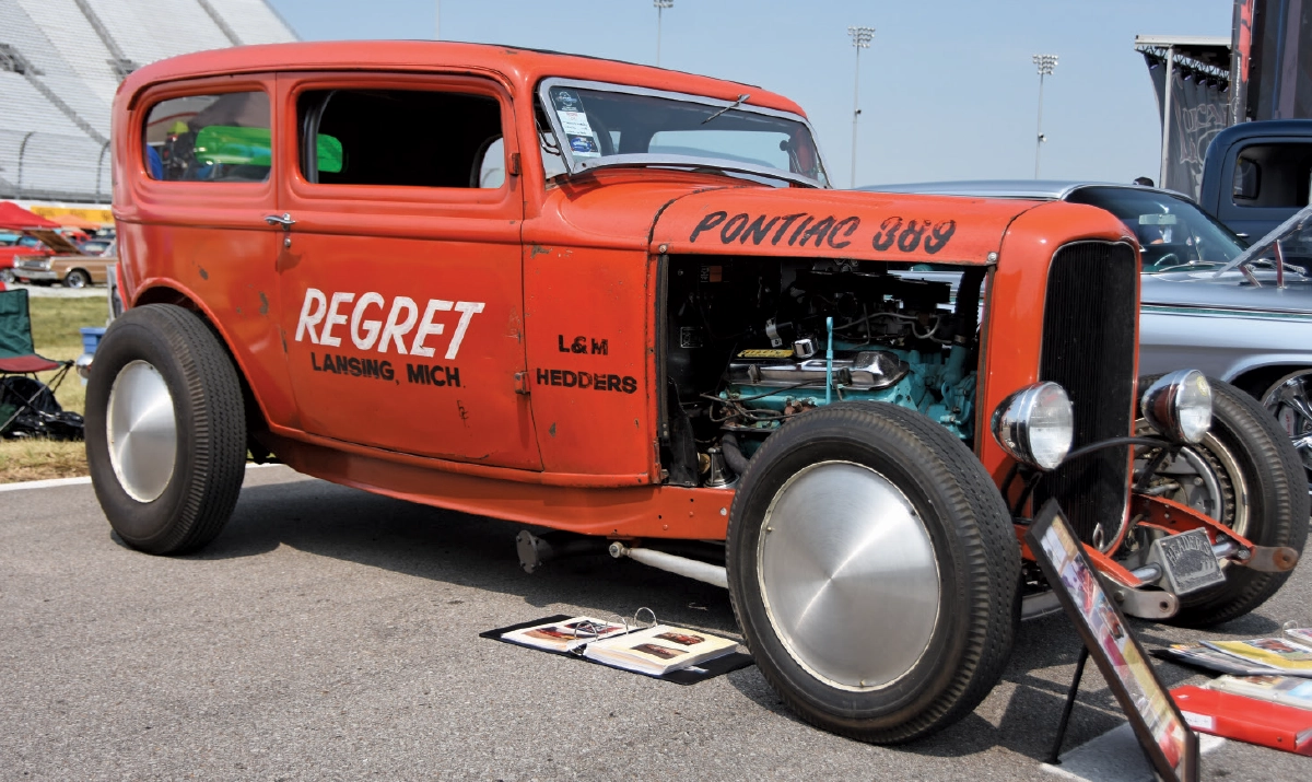 A profile view of the red vintage 1932 vintage Ford hot rod sedan, parked outdoors at a Triple Crown of Rodding car show event, showing the exposed engine and silver disc wheels