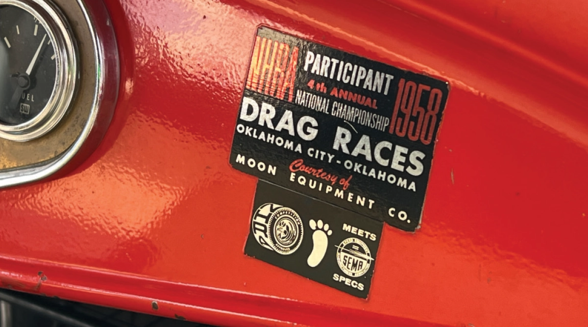 A close-up shot of a red 1932 vintage Ford hot rod sedan's dashboard area featuring a black rectangular sticker that reads PARTICIPANT 4th ANNUAL NATIONAL CHAMPIONSHIP 1958 DRAG RACES OKLAHOMA CITY - OKLAHOMA Courtesy of MOON EQUIPMENT CO.