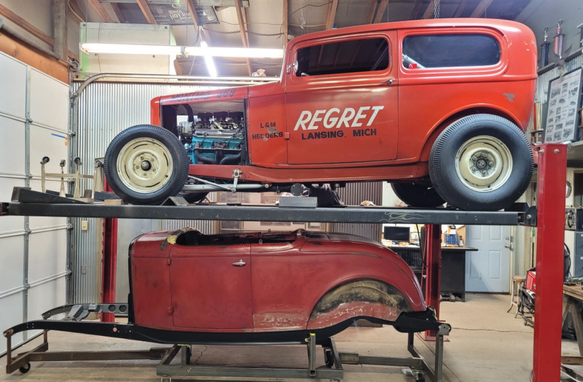An indoor garage scene showing the red vintage 1932 Ford hot rod sedan REGRET on a car lift; Directly beneath the lift sits a separate red car body on a wheeled metal frame