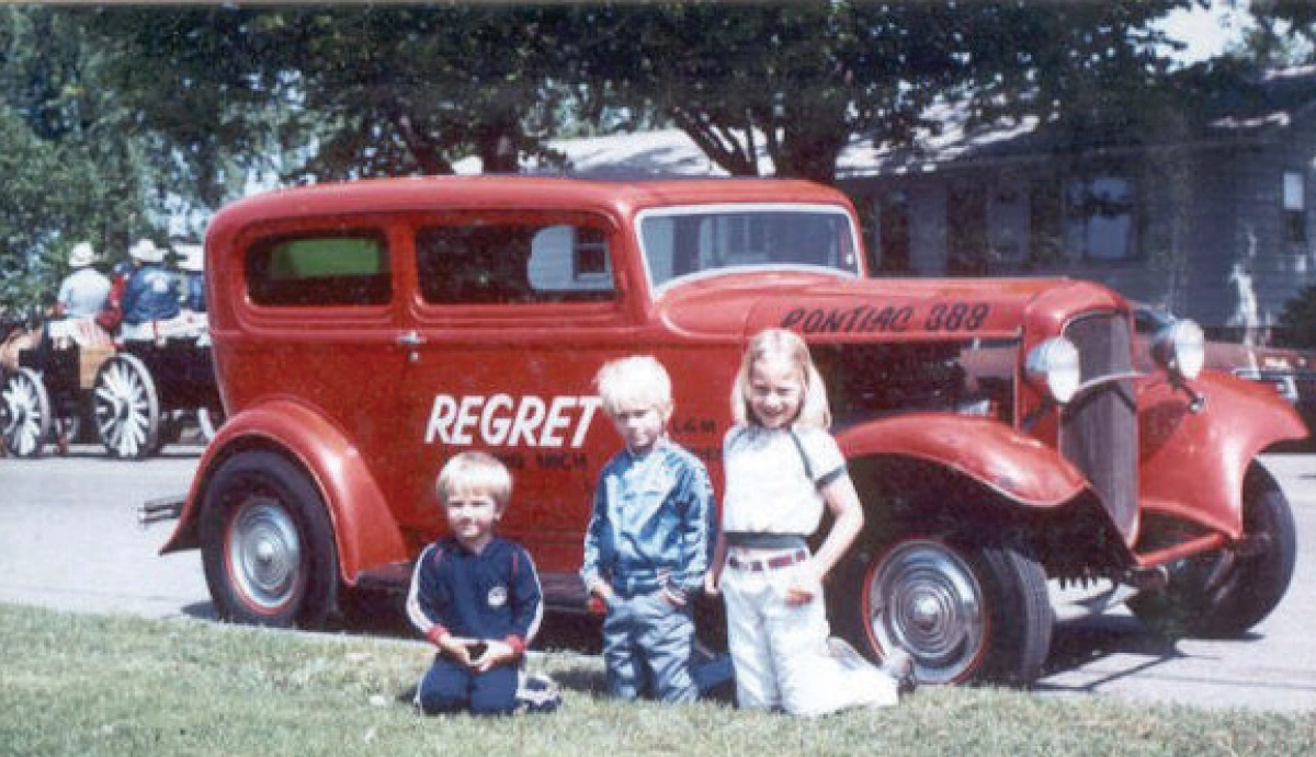 A vintage color photograph of three young children—two boys (Matt and Mike) and a girl (Dawn)—who happen to be Dennis 'Denny' Lesky's kids kneeling in the grass in front of the red vintage 1932 Ford hot rod sedan REGRET