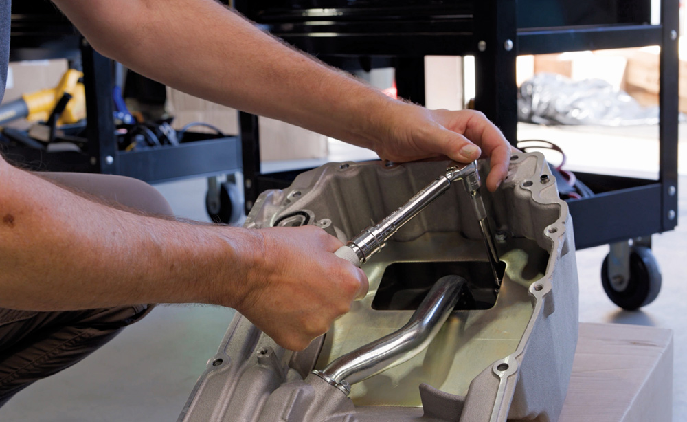 Close-up of a mechanic's hands using a socket wrench to install an internal oil baffle inside a cast aluminum oil pan, ensuring proper oil control for the engine.