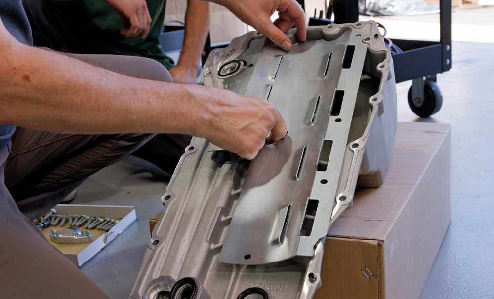 A technician manually positions a long, slotted metal windage tray into a silver aluminum oil pan, preparing the component for installation onto the engine block.