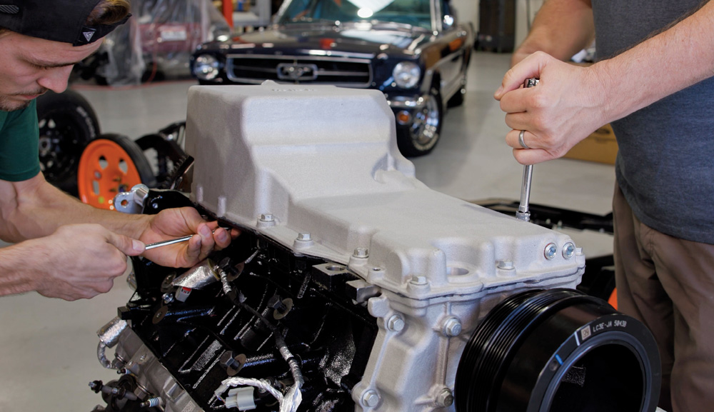 Two mechanics work together to bolt a high-clearance aluminum oil pan onto the bottom of a black engine block using a socket wrench in a well-lit automotive shop.