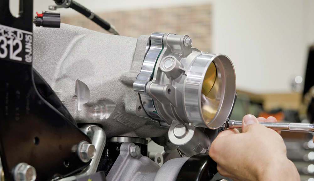 A technician uses a hand tool to bolt a silver high-performance throttle body onto the adapter plate of the aluminum intake manifold on a Godzilla V8 engine.