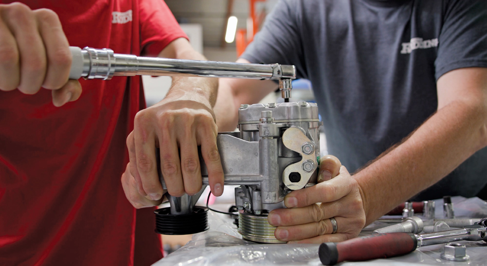 Two mechanics work together to assemble a silver air conditioning compressor on a workbench, using a torque wrench to ensure all fasteners are to specification.