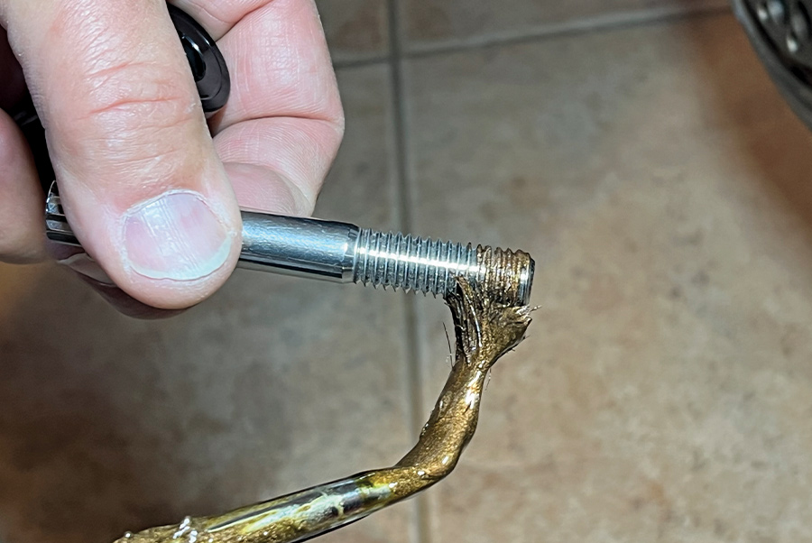 A close-up photograph of a mechanic applying a thick, bronze-colored anti-seize lubricant to the threads of a silver mounting bolt using a small brush to ensure proper torque and prevent galling