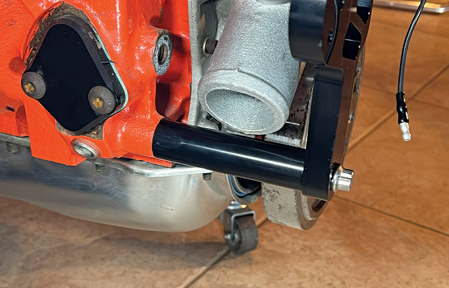 A close-up shot showing a long black anodized spacer and high-strength silver bolt securing the lower engine accessory mounting bracket to an orange V8 engine block