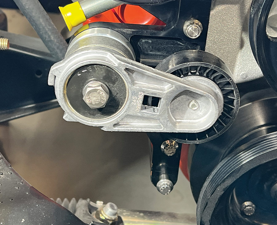 A close-up view of a spring-loaded serpentine belt tensioner fully installed on a black Front Runner mounting bracket, featuring a silver cast-aluminum arm and a black composite pulley