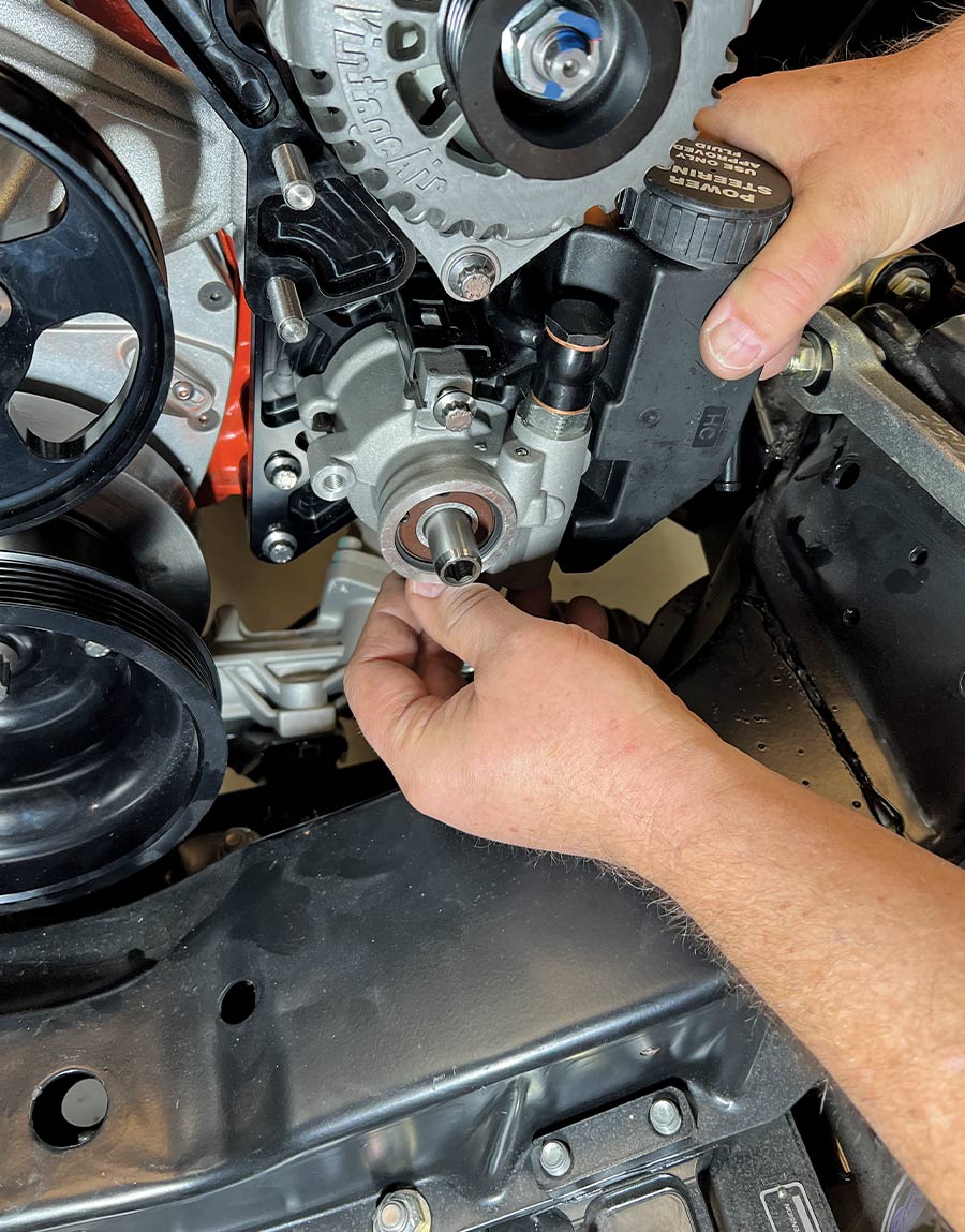 A close-up action shot of a mechanic installing a high-performance power steering pump with an attached black fluid reservoir onto a Front Runner engine accessory drive bracket