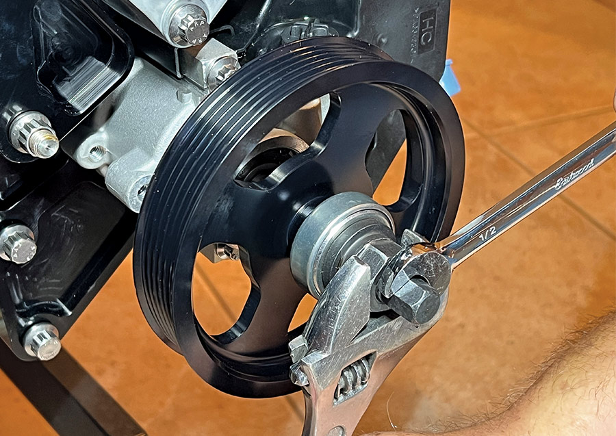 A close-up photograph showing the installation of a black anodized power steering pulley onto a pump shaft using a specialized pulley installer tool, a combination wrench, and an adjustable wrench