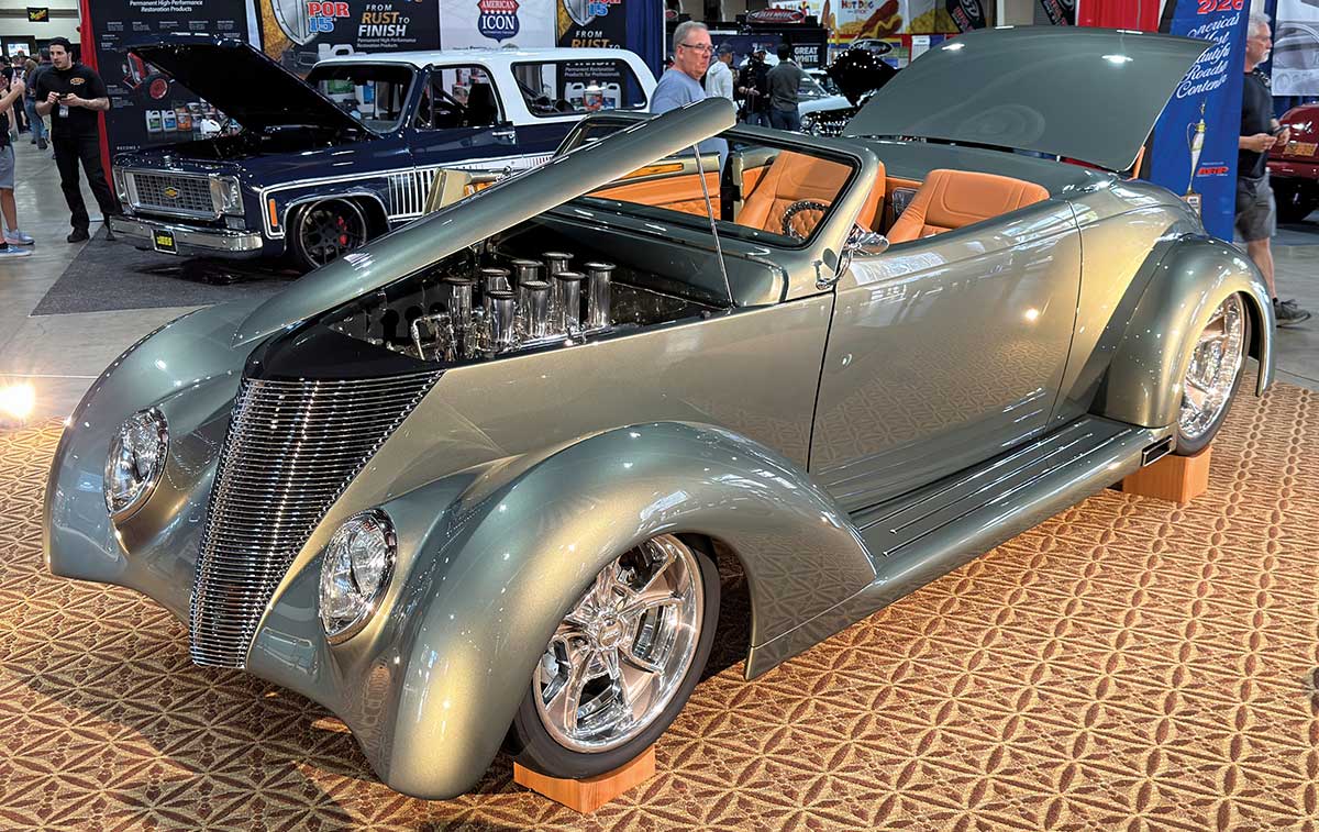 1934 Ford Roadster custom build metallic gray paint, long hood with rounded fenders, low profile tires, open top, tan interior, chrome accents, visible engine components, displayed at an indoor automotive show