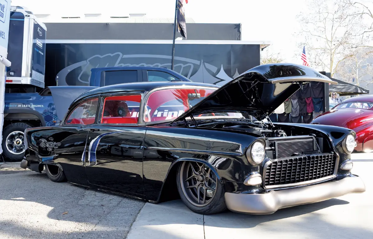 A low-profile, Ryno Built 1955 Chevrolet Bel Air black car displayed outdoors on a sunny day parked near large event trailers and other show cars