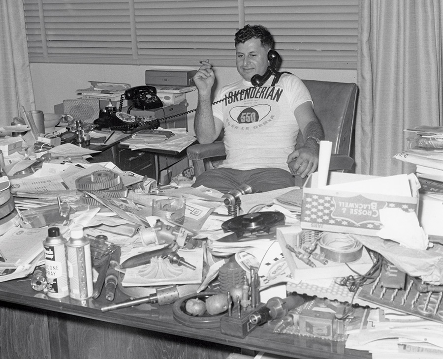 Black and white photograph of Isky sitting at a desk heavily cluttered with automotive parts, papers, and spray cans. He wears an Iskenderian t-shirt, holds a cigar in one hand, and has a rotary telephone handset to his ear.