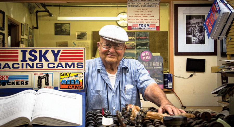 Color photo of Isky in a white flat cap and blue striped shirt smiling behind an Isky Racing Cams counter. He rests his hand on metal engine parts near an open catalog. Customer service signage and framed photos are in the background.