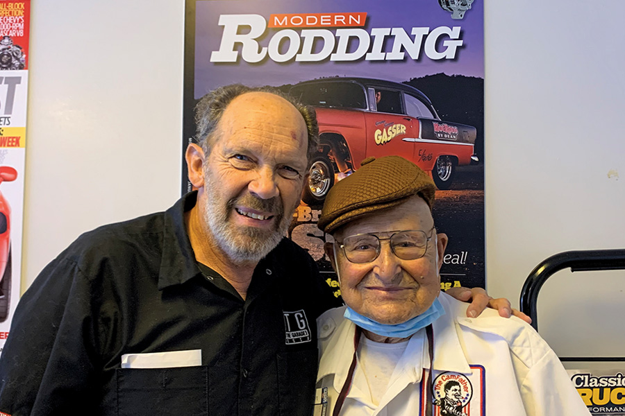 Color photo of Brian Brennan and Ed “Isky” Iskenderian in front of a Modern Rodding poster. Brian Brennan stands on the left wearing a black button-down shirt, on the right Ed wears a brown flat cap and a white shirt with a “The Camfather” patch.