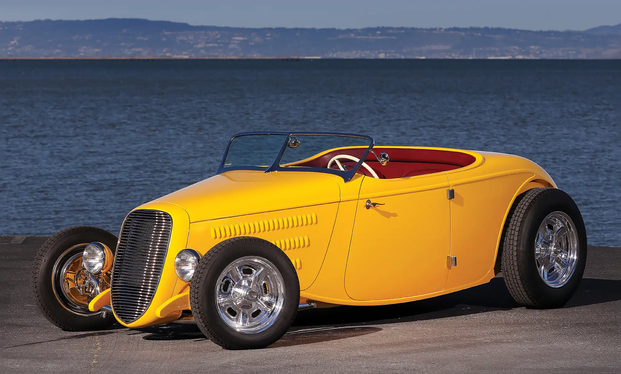A bright yellow vintage hot rod roadster with a red interior parked next to a large body of blue water with distant hills on the horizon.