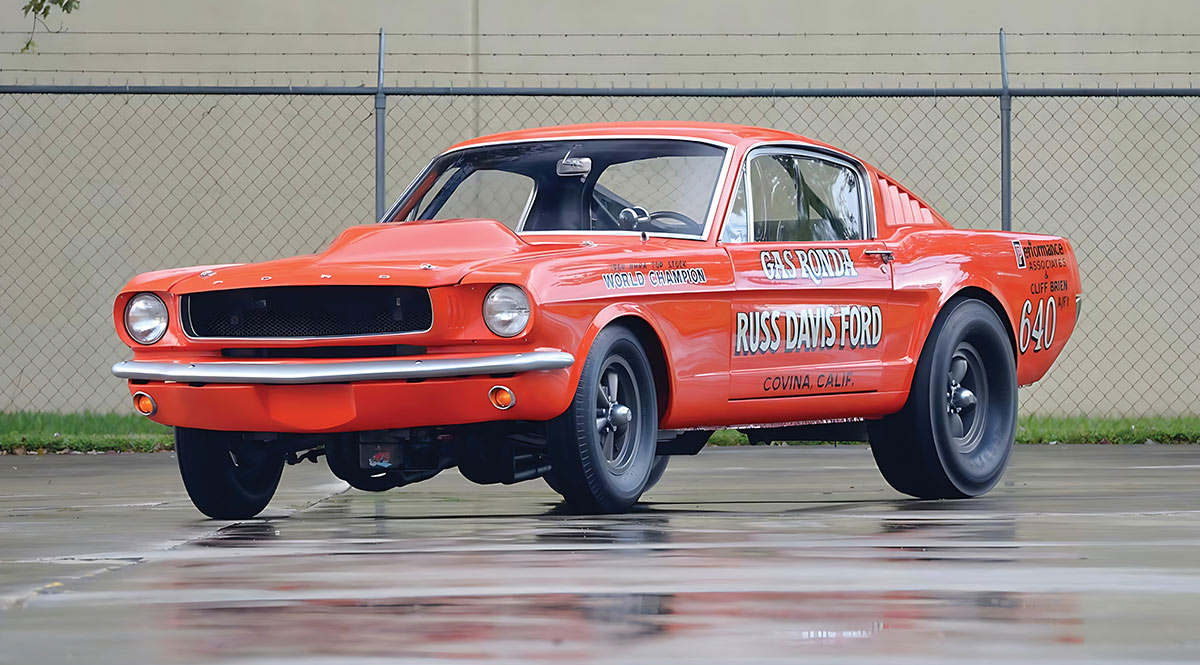 A classic orange Ford Mustang fastback drag car with "Gas Ronda" and "Russ Davis Ford" lettering, parked on a wet, reflective surface in front of a chain-link fence.