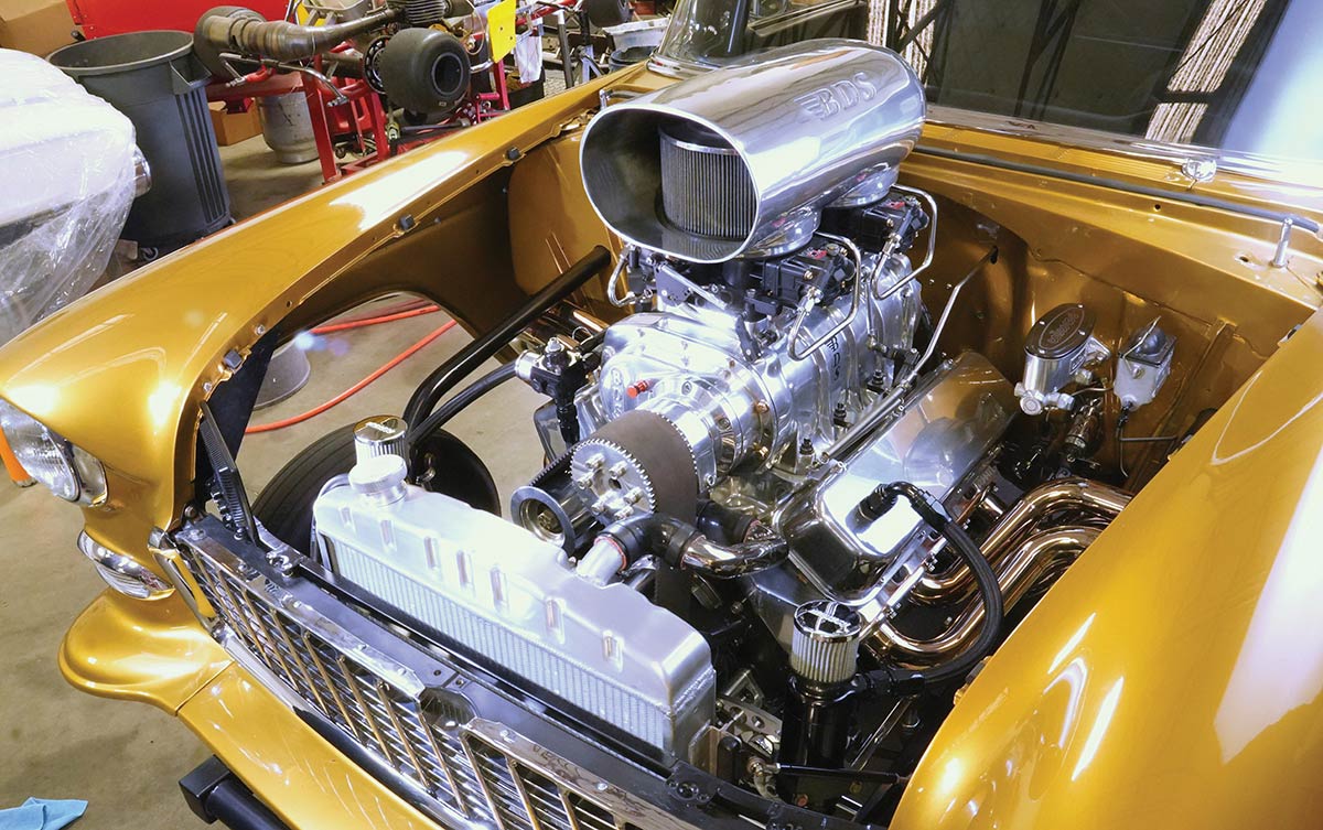 Engine bay of a 1955 Chevy Bel Air featuring a supercharged V8 engine.