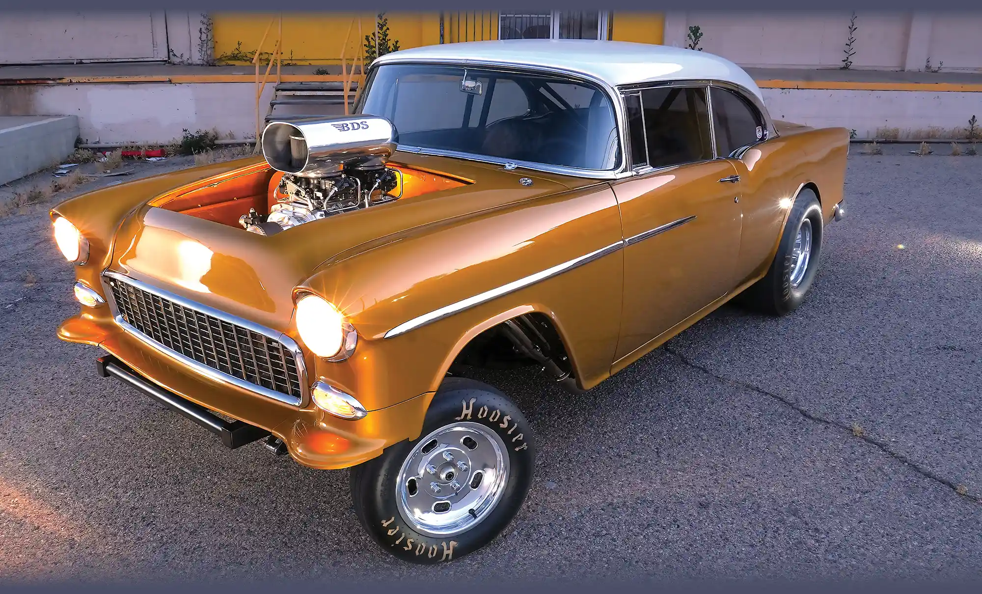 Gold and white 1955 Chevrolet Bel Air gasser with headlights on, parked outdoors.