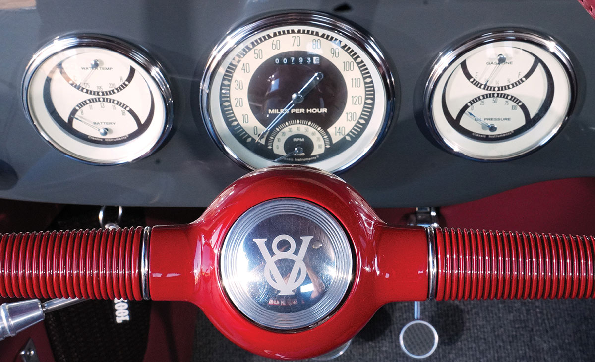 Vintage hot rod dashboard featuring three chrome-rimmed Classic Instruments gauges behind a red steering wheel with a chrome Ford V8 horn cap.